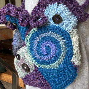 May include: A handmade crochet bag in shades of blue, purple, and gray. The bag features a spiral design and is adorned with large, dark brown buttons. The strap is also crocheted, and the bag appears to be worn over the shoulder.