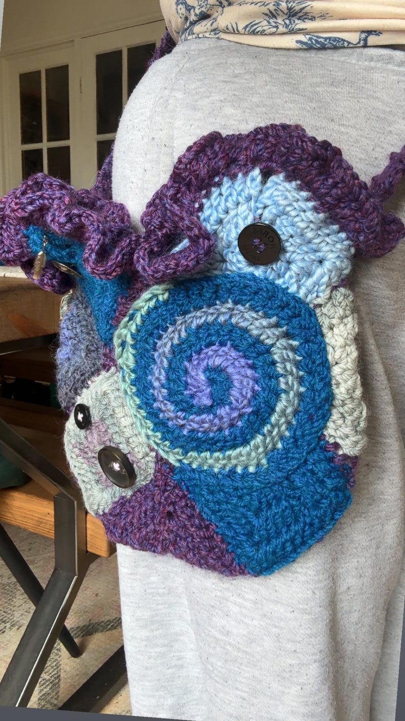 May include: A handmade crochet bag in shades of blue, purple, and gray. The bag features a spiral design and is adorned with large, dark brown buttons. The strap is also crocheted, and the bag appears to be worn over the shoulder.