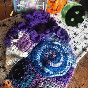 May include: A handmade crochet bag in shades of purple, blue, and white, featuring a spiral design and two large black buttons. The bag is displayed with a Halloween-themed wallet, a carved pumpkin, and a decorative moon.