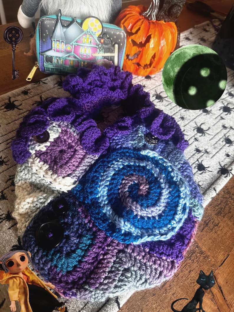May include: A handmade crochet bag in shades of purple, blue, and white, featuring a spiral design and two large black buttons. The bag is displayed with a Halloween-themed wallet, a carved pumpkin, and a decorative moon.