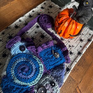 May include: A handmade crochet bag in shades of blue, purple, and gray, featuring a spiral design and decorative buttons. The bag has a purple strap and is placed on a spider-themed runner, with a carved orange pumpkin and a black cat figurine nearby.