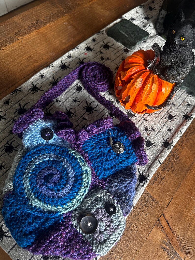 May include: A handmade crochet bag in shades of blue, purple, and gray, featuring a spiral design and decorative buttons. The bag has a purple strap and is placed on a spider-themed runner, with a carved orange pumpkin and a black cat figurine nearby.