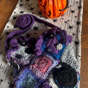 May include: A handmade crochet bag in shades of purple, blue, gray, and pink, featuring a patchwork design and black buttons. The bag has a long strap and a decorative, rolled top. An orange carved pumpkin sits in the background.