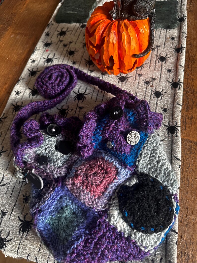 May include: A handmade crochet bag in shades of purple, blue, gray, and pink, featuring a patchwork design and black buttons. The bag has a long strap and a decorative, rolled top. An orange carved pumpkin sits in the background.