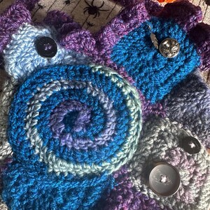 May include: A handmade crochet bag in shades of blue, purple, and gray. The bag features a spiral design and is adorned with decorative buttons. The strap and edges are a darker purple.