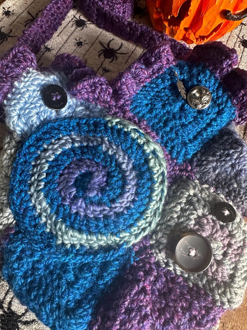 May include: A handmade crochet bag in shades of blue, purple, and gray. The bag features a spiral design and is adorned with decorative buttons. The strap and edges are a darker purple.