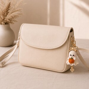 Peut inclure: Un sac &agrave; bandouli&egrave;re couleur cr&egrave;me avec une finition lisse et une fermeture &agrave; rabat. Attach&eacute; &agrave; la boucle m&eacute;tallique lat&eacute;rale se trouve un petit fant&ocirc;me en crochet tenant une minuscule citrouille orange, avec un n&oelig;ud et une feuille.