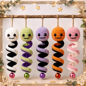 May include: Five crocheted hanging ornaments featuring round heads with black eyes and stitched mouths, each attached to a spiral-shaped body in white, green, purple, orange, or pink, with a small metal bell dangling from the bottom of each.