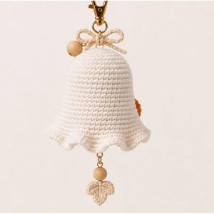 Peut inclure: Une cloche d&eacute;corative en crochet de couleur cr&egrave;me avec une petite perle en bois et un n&oelig;ud en crochet sur le dessus. Un fermoir mousqueton en m&eacute;tal dor&eacute; est fix&eacute; au sommet. Une perle en bois et une petite feuille en crochet pendent au centre.