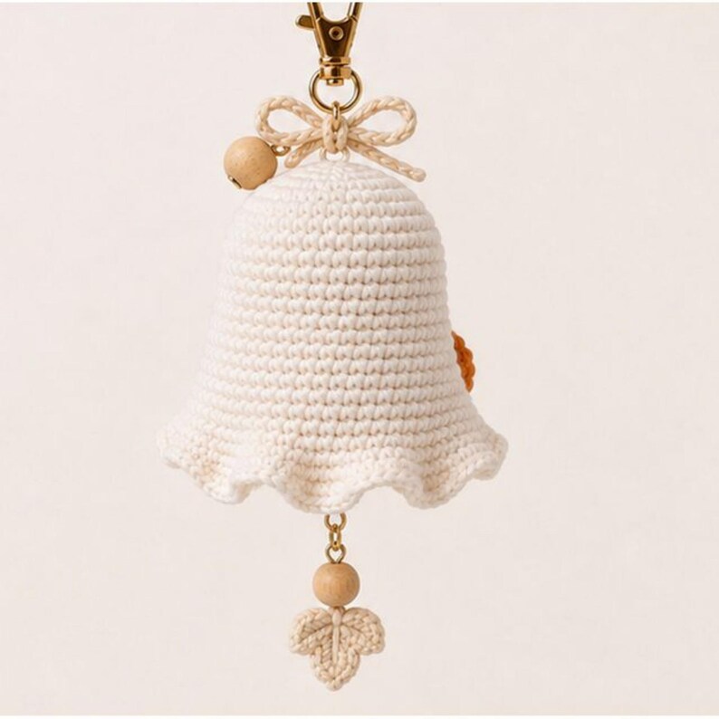Peut inclure: Une cloche d&eacute;corative en crochet de couleur cr&egrave;me avec une petite perle en bois et un n&oelig;ud en crochet sur le dessus. Un fermoir mousqueton en m&eacute;tal dor&eacute; est fix&eacute; au sommet. Une perle en bois et une petite feuille en crochet pendent au centre.