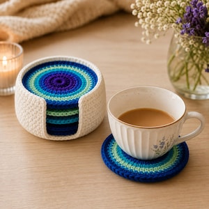 May include: A set of round, crocheted coasters featuring concentric rings in shades of dark blue, bright blue, and light green. The coasters are stored in a matching white, circular, crocheted holder, with one coaster placed under a white ceramic mug.
