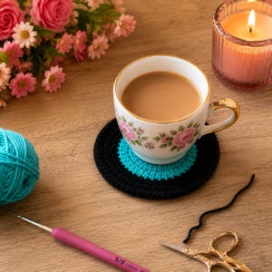 May include: A round, two-toned crochet coaster featuring a black outer ring and a bright turquoise inner circle. The coaster supports a white ceramic teacup filled with light brown tea, decorated with pink rose patterns and gold trim.