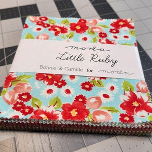 May include: A stack of fabric squares with a floral pattern in red, white, and green on a light blue background. The fabric is wrapped with a white label that reads "moda Little Ruby Bonnie & Camille for moda."