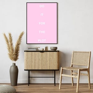 Könnte beinhalten: Ein rosa gerahmtes Poster mit dem Text "DO IT FOR THE PLOT" in weißen Buchstaben. Das Poster hängt an einer weißen Wand über einem hellbraunen Schrank mit schwarzem Metallrahmen. Ein hellbrauner Korbstuhl steht im Vordergrund.