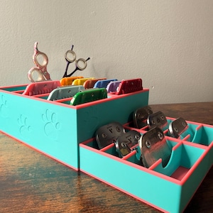 May include: A tiered, turquoise organizer with pink trim, holding grooming tools. The organizer features paw print cutouts and contains various clipper blades, scissors, and colored comb attachments. The tools are arranged in separate compartments.