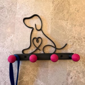 Può includere: Porta guinzaglio per cani in metallo nero con silhouette di cane e pomelli rosa. La silhouette del cane ha un cuore al centro. Un guinzaglio blu è appeso a uno dei pomelli. Il supporto è montato su una parete chiara.