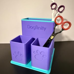Peut inclure: Un organisateur de bureau violet et bleu sarcelle à trois compartiments, avec le texte "Dogfinity" en relief et des motifs de pattes sur le devant. Une paire de ciseaux de toilettage aux anneaux violets et orange repose dans le compartiment le plus haut.