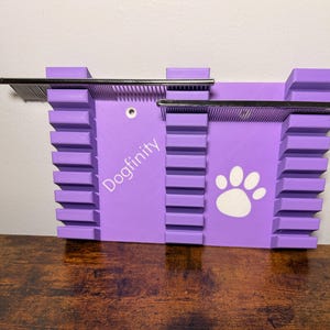 Puede incluir: Un organizador de pared morado con tres columnas verticales con ranuras horizontales, dos barras de metal plateado, el texto Dogfinity y un gráfico de huella blanca. La unidad incluye dos orificios de montaje circulares para instalar.