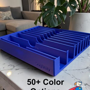 Op de afbeelding: Een blauwe rechthoekige organizerbak met meerdere parallelle verdelers en de in reliëf aangebrachte tekst Dogfinity op de voorrand. De bak bevat de gedrukte tekst 50+ Color Options en een afbeelding van een hond met een rode cape.