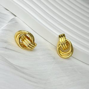 Peut inclure: Une paire de boucles d'oreilles dorées, chacune composée de trois anneaux circulaires entrelacés avec une finition polie et réfléchissante. Les boucles d'oreilles présentent une structure métallique lisse et arrondie, présentées en paire.