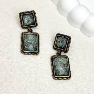 May include: A pair of vintage-style earrings. Each earring has two square bronze-toned frames, connected by a small loop. The frames have a textured, rope-like border and contain a light green, fibrous material. The earrings are displayed on a white surface.