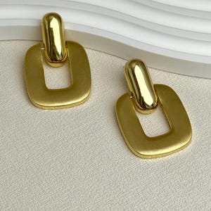 May include: A pair of gold-colored earrings. Each earring features a rectangular open shape with a rounded top and a connecting bar. The earrings have a matte finish and are displayed on a textured surface.