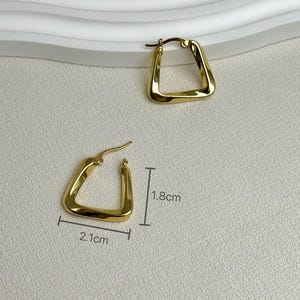 May include: A pair of gold-toned hoop earrings with a rounded triangular shape. One earring is shown with measurement lines indicating a width of 2.1 cm and a height of 1.8 cm. The earrings feature a smooth, polished finish and a hinged post closure.