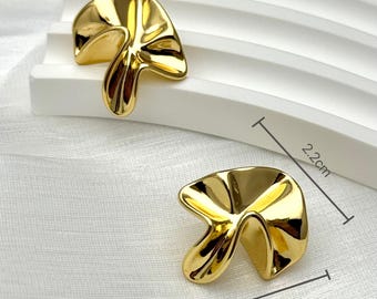 May include: Two gold-colored, abstract-shaped earrings. Each earring has a unique, flowing design. The earrings are approximately 2.5 cm wide and 2.2 cm high. They are displayed on a white surface with a textured background.