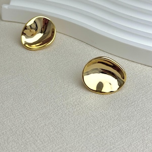 May include: A pair of gold-toned stud earrings with a smooth, reflective, and irregular circular shape. The earrings feature a concave center and a polished metallic finish that catches the light, creating subtle highlights across the curved surface.
