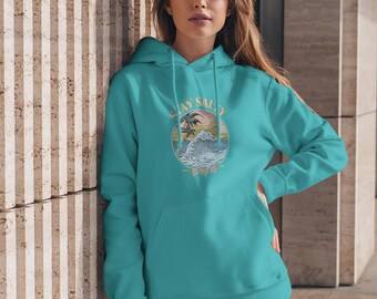 Puede incluir: Sudadera con capucha turquesa con un diseño gráfico de una ola, palmeras y las palabras "Stay Salty" en un círculo. La sudadera tiene una capucha con cordón y un bolsillo delantero. La persona lleva vaqueros azules.