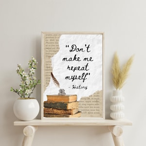 May include: A framed print with the quote "Don't make me repeat myself" and the word "History" below it. The print features a stack of old books, a quill pen, and a small inkwell. A white vase with flowers and a decorative vase with dried grass are on either side.