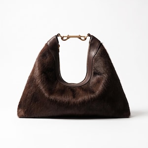 May include: A dark brown hobo bag crafted from textured hair-on hide, featuring a smooth dark brown leather trim along the top opening and a central metallic gold-toned hardware clasp connecting the two sides of the handle structure.