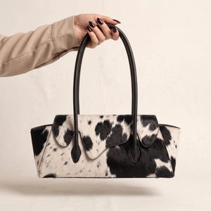 May include: A rectangular handbag featuring a black and white cow print pattern with a textured hair-on-hide finish. The bag includes two tall, slim black leather handles and a flap closure with an asymmetrical, scalloped edge design.