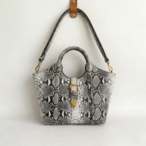 Könnte beinhalten: Eine Handtasche mit grauem und weißem Schlangenmuster, mit einem oberen Griff und einem langen Schulterriemen. Die Tasche hat einen goldfarbenen Schnallenverschluss und ist an einem Lederriemen aufgehängt.