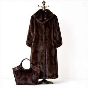May include: A full-length brown faux fur coat with a wide collar hangs on a brass clothing rack. A matching brown handbag with leather trim sits at the base of the rack. The coat has long sleeves and a button closure.