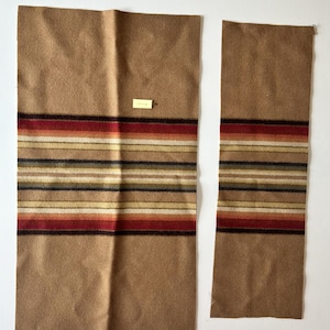 May include: Two rectangular pieces of tan fabric featuring a horizontal striped pattern in shades of red, orange, yellow, and dark brown. The larger piece includes a small, rectangular yellow tag attached to the center of the fabric surface.