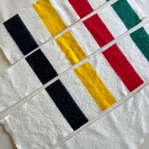 May include: Close-up of several white fabric strips with bold, diagonal stripes in black, yellow, red, and green. The strips have a soft, textured appearance, suggesting a towel or similar textile. The design is simple and colorful.