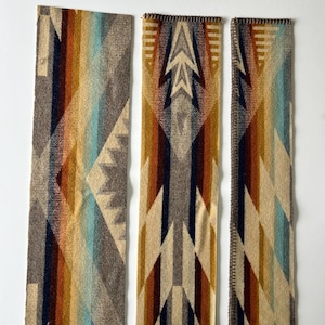 May include: Three rectangular fabric strips featuring a woven geometric pattern in shades of tan, brown, dark blue, and light blue. The strips display repeating angular shapes and chevron motifs, with one strip showing a visible dark stitched edge.