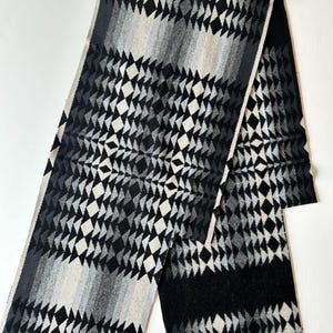 May include: A long rectangular scarf featuring a repeating geometric pattern of diamonds and triangles in a gradient of black, charcoal gray, light gray, and white. The woven fabric displays a symmetrical design with alternating dark and light sections.