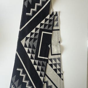 May include: A black and white geometric patterned scarf. The scarf features a repeating pattern of triangles, zigzags, and diagonal lines. The scarf is made of a soft material and is likely for cold weather.
