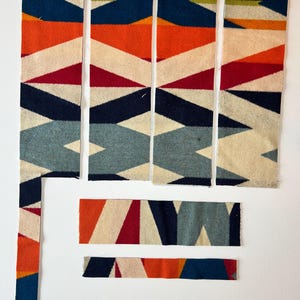May include: Six rectangular fabric strips and one long vertical strip featuring a geometric pattern in shades of navy blue, orange, red, cream, and light blue. The fabric has a woven texture with repeating triangular and diamond shapes.
