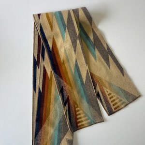 May include: A rectangular scarf with a geometric pattern in shades of tan, brown, blue, and orange. The scarf features a repeating diamond design and is made of a soft, woven material.