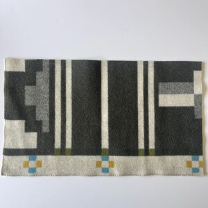 May include: A rectangular, patterned textile featuring a geometric design in shades of gray, white, and touches of blue and yellow. Vertical white stripes and square accents create a structured look. The textile is likely a scarf or blanket.