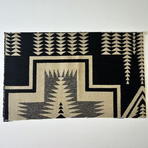 May include: A rectangular fabric swatch featuring a high-contrast black and beige geometric pattern. The design includes repeating rows of small triangles and a large, central stepped motif with jagged, saw-tooth edges throughout the textile.