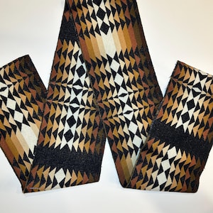 May include: A long, rectangular fabric strip featuring a repeating geometric pattern of triangles and diamonds. The design incorporates a color palette of black, cream, tan, and various shades of brown, arranged in a symmetrical, interlocking layout.