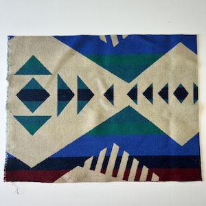 May include: A rectangular woven textile featuring a geometric pattern with large beige diamonds, blue and teal triangles, and horizontal bands of dark blue and deep burgundy. The fabric displays a repeating sequence of sharp angular shapes.