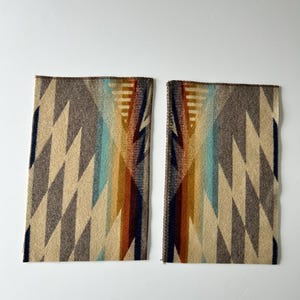 May include: Two rectangular fabric swatches featuring a geometric pattern with repeating diamond shapes in shades of beige, brown, dark blue, light blue, and orange. The edges of the fabric are finished with a dark brown stitched border.