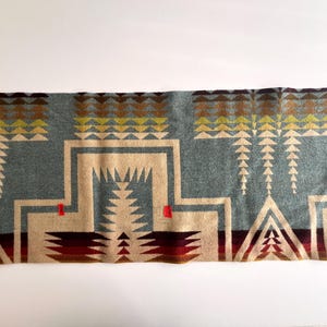 May include: A rectangular blanket with a geometric pattern in shades of blue, beige, brown, and red. The design features repeating triangular and rectangular shapes. The blanket has a brown border and is likely made of wool or a similar material.
