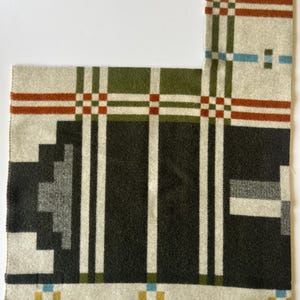 May include: A rectangular blanket with a geometric pattern. The blanket features a dark green and black central section, bordered by cream-colored sections with red, green, and blue accents. White vertical stripes run through the design.