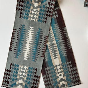 May include: A folded textile with a geometric pattern in shades of gray, teal, brown, and cream. The design features repeating triangular shapes and a textured appearance. The edges are unfinished, revealing the fabric's weave.